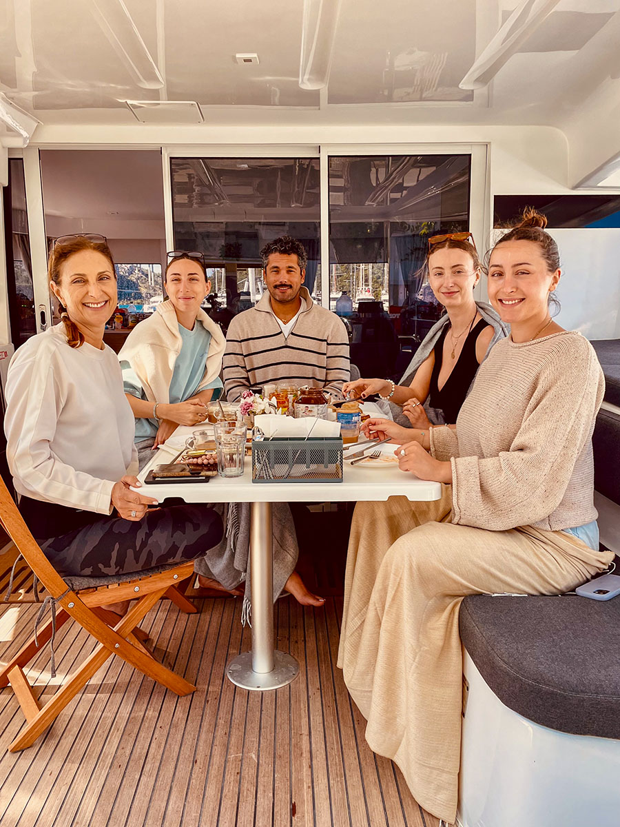 Five guests smiling around the breakfast table in the catamaran cockpit
